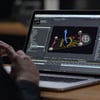 A person uses a laptop displaying 3D animation software focused on a skateboard model with directional arrows.